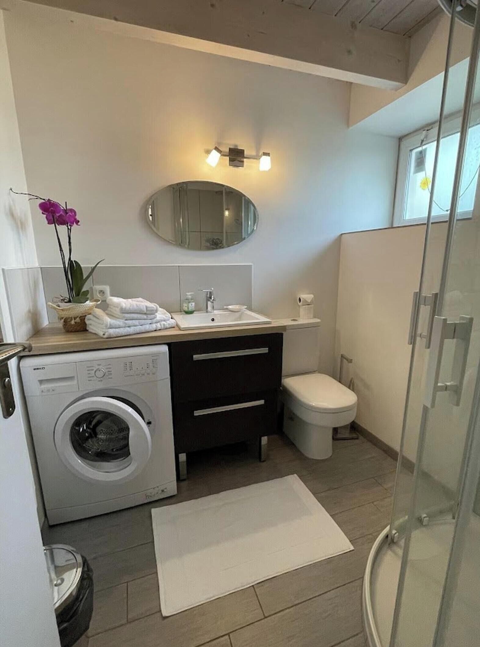 Une salle de bain moderne équipée d’un lave-linge, de toilettes, d’une douche à parois de verre et d’un miroir avec des appliques murales.