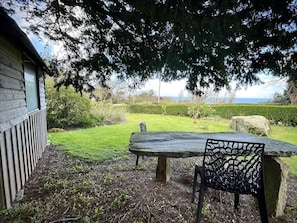 A stone table and chair in a garden with a wooden building and a view of the sea. - Rental for 2 With Private Outdoor Space (Plougasnou)