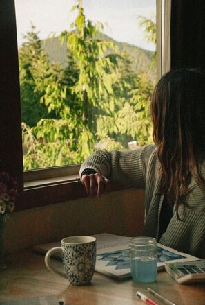 Une personne appuyée sur le rebord d’une fenêtre, avec une tasse et un verre posés sur la table.