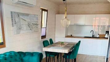 A dining area with a wooden table and chairs, a sofa, a kitchen with white cabinets, and a hanging light fixture.