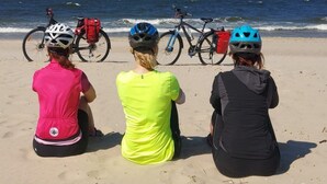 Tre personer sidder på en sandstrand med cykler og hjelme, vendt mod havet.