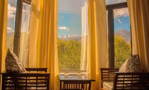 A room with a table and chairs, a view of mountains and greenery through large windows with curtains. - Hotel Nature Residency Leh Ladakh (Leh)