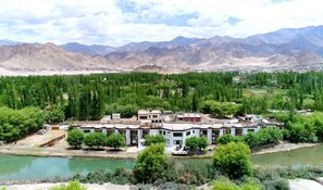 A building with multiple rooms, a river in the foreground, and mountains in the background. - Hotel Nature Residency Leh Ladakh (Leh)
