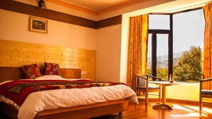A bedroom with a wooden headboard, a bed with a white comforter, a small table with a chair, and a window with curtains. - Hotel Nature Residency Leh Ladakh (Leh)