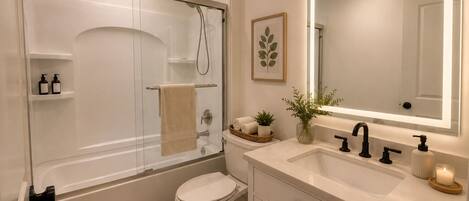Un baño moderno con ducha de vidrio, inodoro, lavamanos con espejo y una planta pequeña en el mueble.