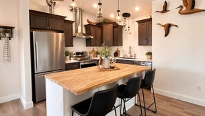 A modern kitchen with a wooden island, stainless steel appliances, hanging pendant lights, and wall-mounted wooden bird sculptures. - Kodiak Suites (Kodiak)