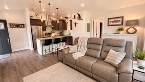 A modern living room with a sofa, a dining area with bar stools, a kitchen with stainless steel appliances, and pendant lighting. - Kodiak Suites (Kodiak)