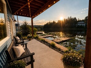 A covered porch with wooden benches, a potted plant, and a view of a lake with lily pads and a dock. - Kodiak Suites (Kodiak)