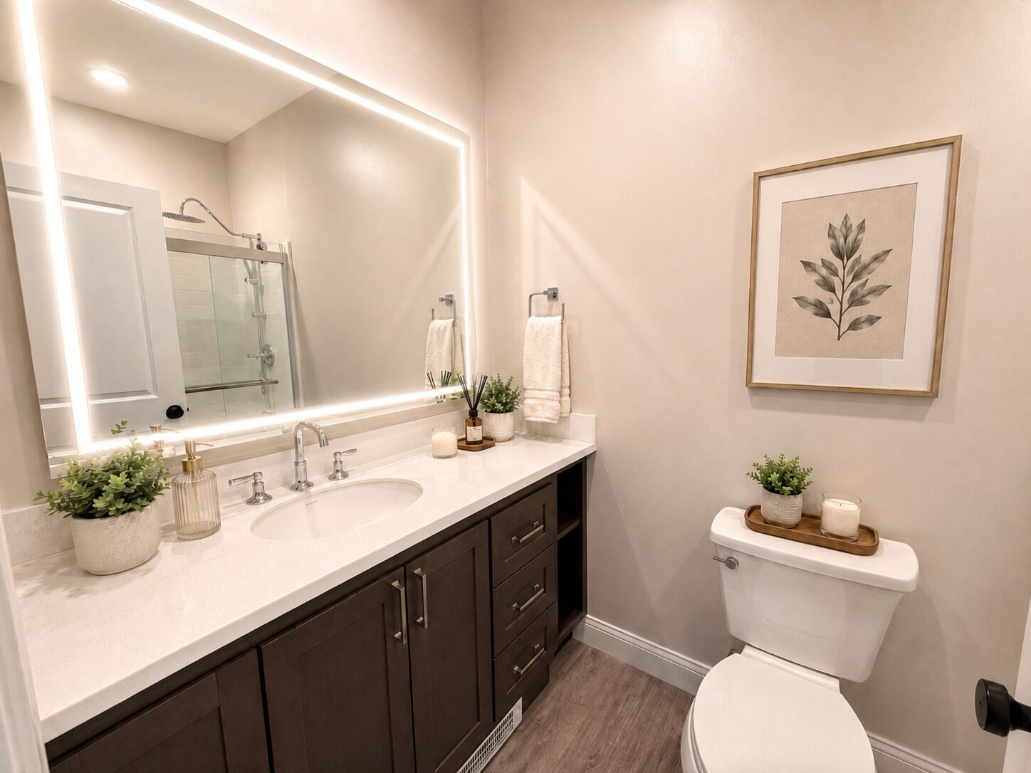 A bathroom with a large mirror, two sinks, dark cabinets, a toilet, and a framed plant on the wall.