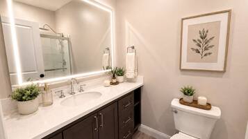 A bathroom with a large mirror, two sinks, dark cabinets, a toilet, and a framed plant on the wall.