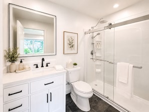 Un baño moderno con tocador blanco, inodoro, ducha con cerramiento de vidrio y un espejo grande.
