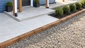 Una casa moderna de dos pisos con un porche cubierto, una puerta principal y un pequeño jardín con plantas en macetas.