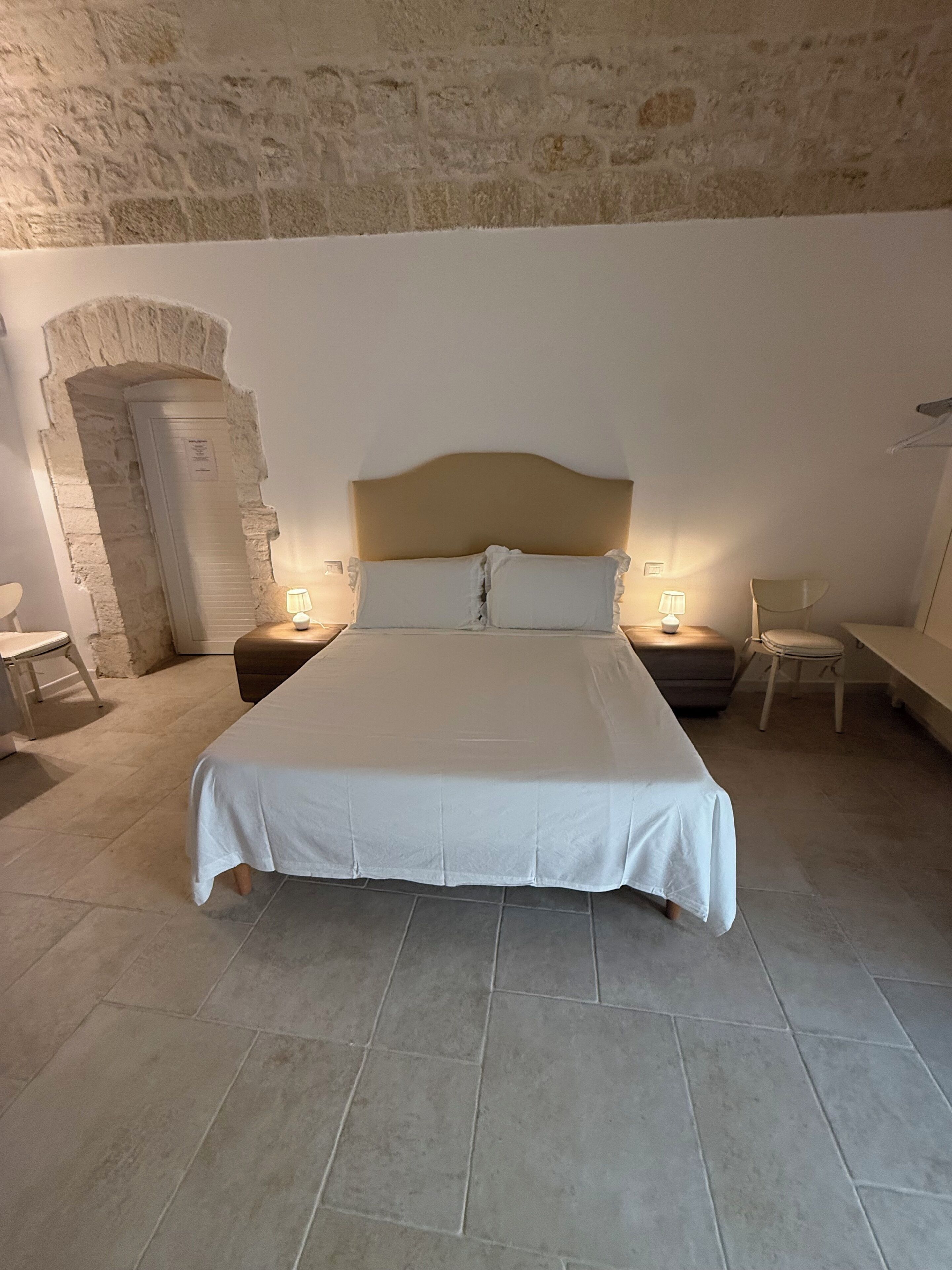 A bedroom with a large bed, two bedside tables, a chair, and a stone wall.