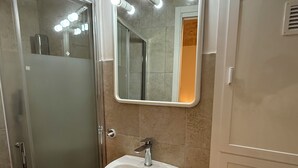 Un baño con cabina de ducha de vidrio, un lavamanos blanco con un espejo arriba y paredes de baldosas color beige.