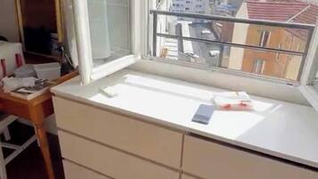 A room with a large window, a desk with drawers, and a view of city buildings.