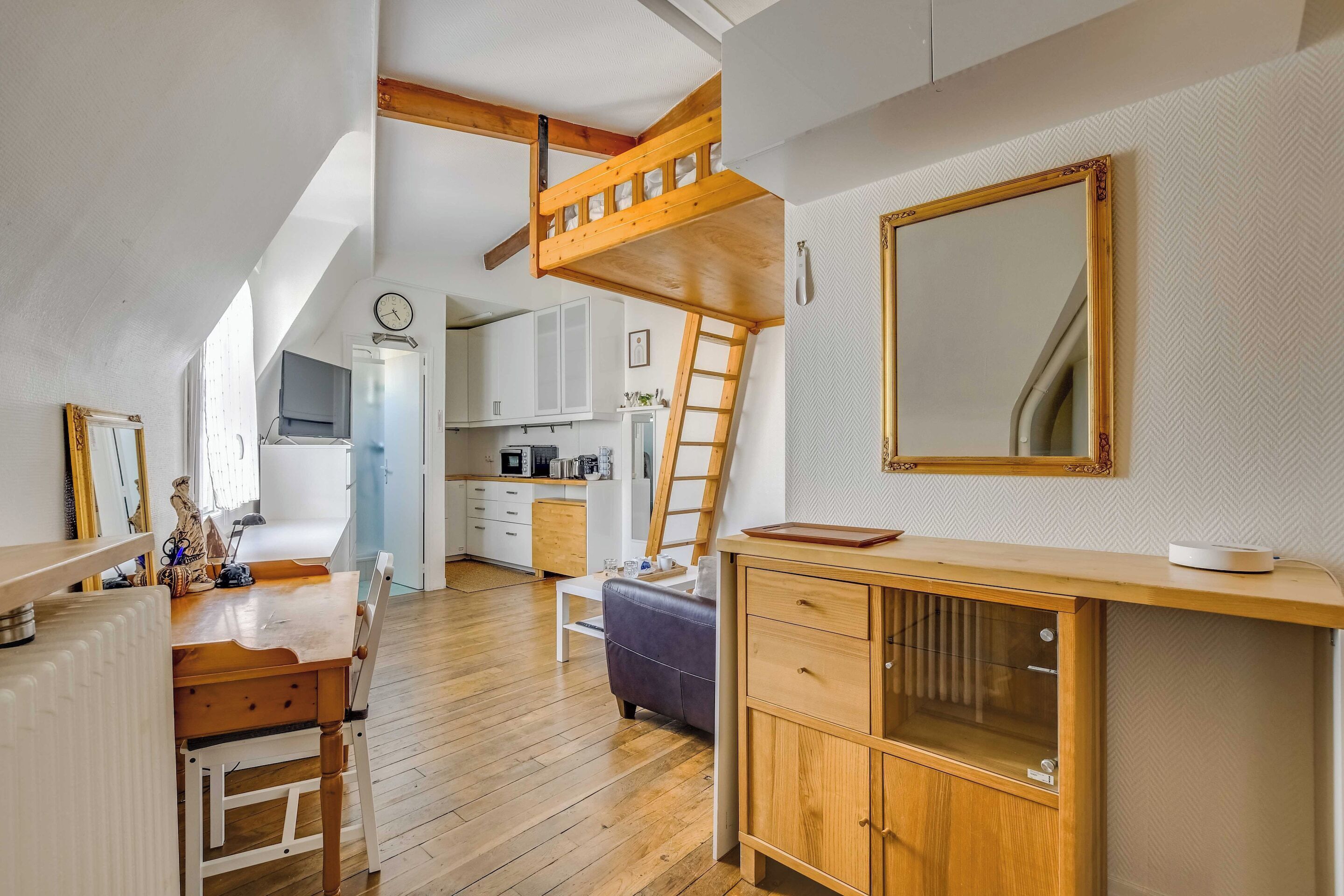 A loft-style room with a wooden loft bed, a dining table with chairs, a kitchen area, and a large mirror.
