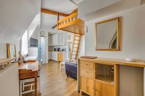 Ein Loft-Zimmer mit einem Schlafplatz in der Zwischengeschoss-Etage, einem Esstisch mit Stühlen, einer Küchenzeile und einem großen Spiegel.