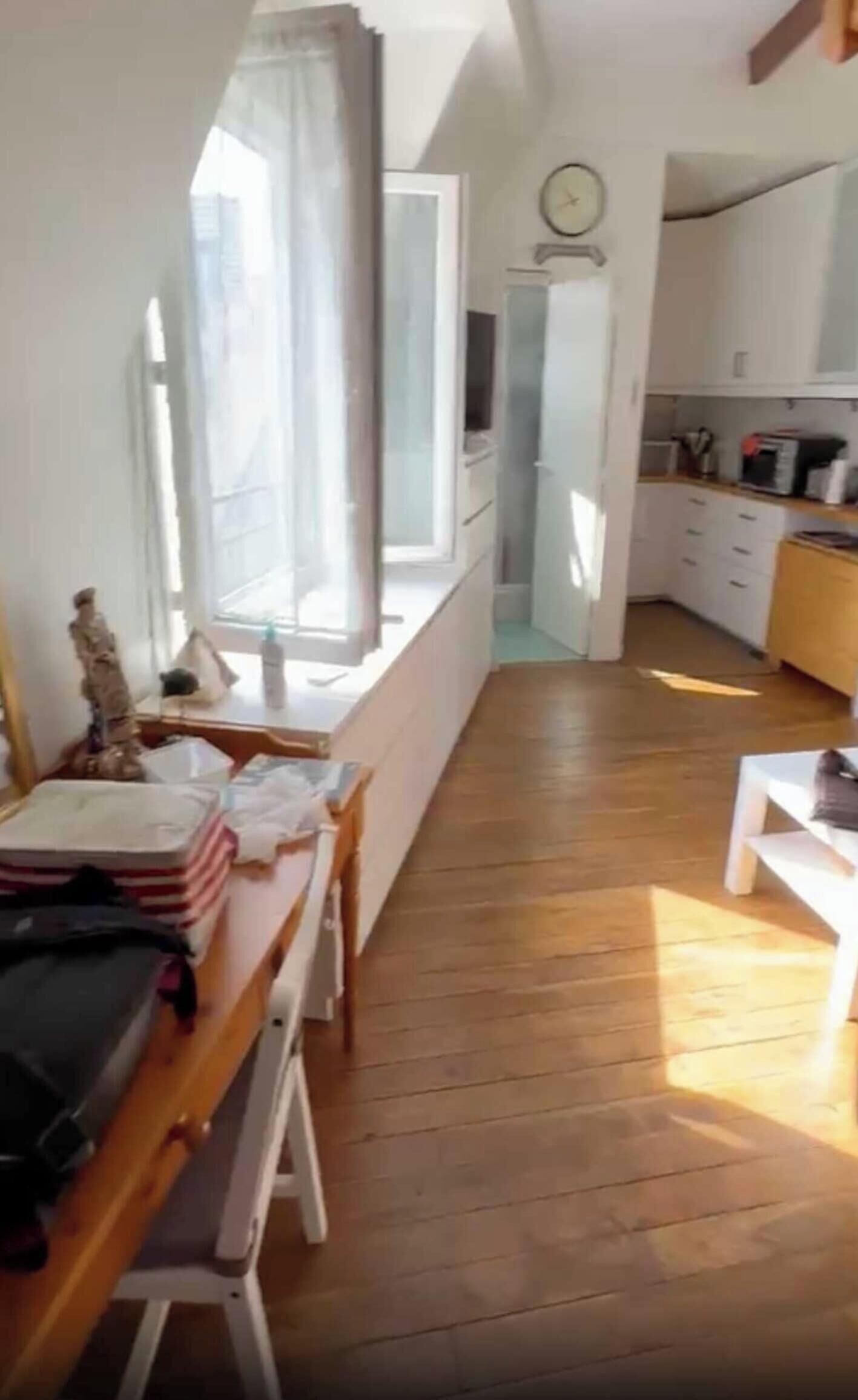 A room with a wooden floor, a dining table with chairs, a window with curtains, a kitchen area with cabinets, and a clock on the wall.