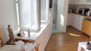 A room with a wooden floor, a dining table with chairs, a window with curtains, a kitchen area with cabinets, and a clock on the wall. - Studio 2P Near Tolbiac / Olympiades (Paris)
