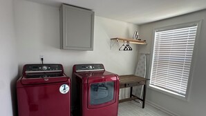 Une buanderie avec deux sèche-linge rouges, une table en bois, une étagère avec des planches à repasser et une fenêtre avec des stores.