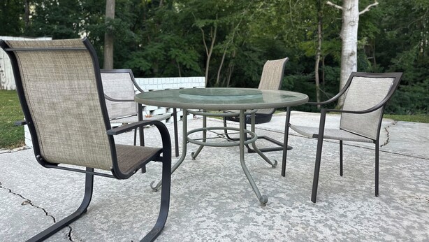 Une terrasse extérieure avec une table ronde en verre et quatre chaises dotées d’une structure en métal et d’un siège en tissu.