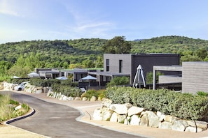 A modern building with a curved driveway, stone landscaping, and a view of a forested hill. - Mediterranean Oasis Near Cala Rossa (Lecci)