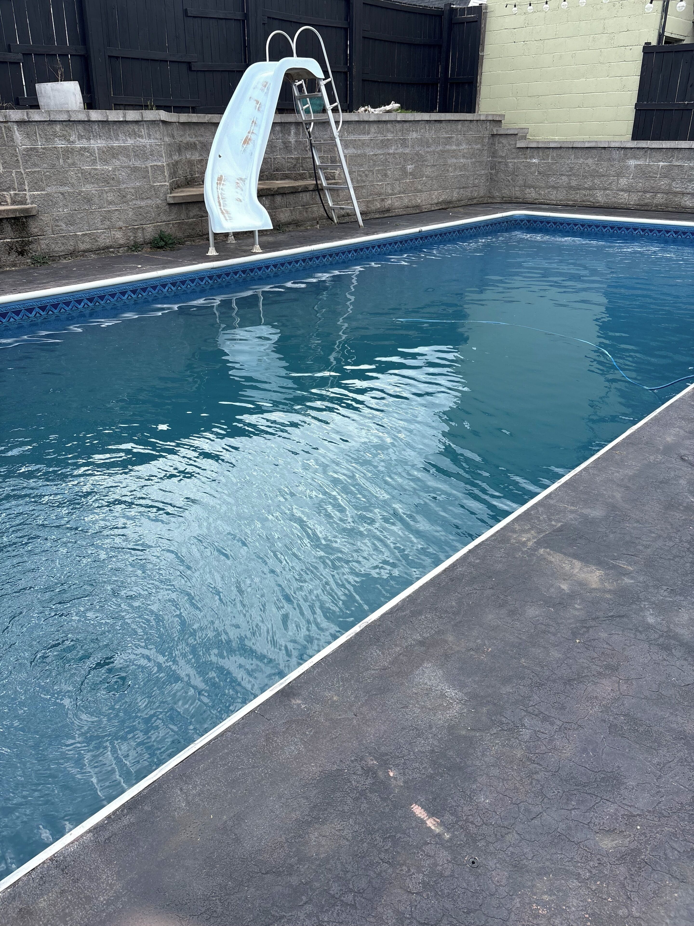 A swimming pool with a ladder, surrounded by a stone wall and a fence.