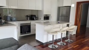 Una cuina moderna amb armaris blancs, una illa central amb tamborets i una zona de menjador amb un sofà gris.