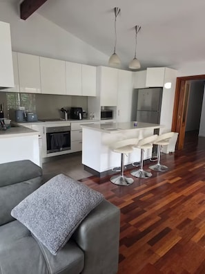 Une cuisine moderne avec des meubles blancs, un îlot central doté de tabourets de bar et un coin repas avec un canapé gris.