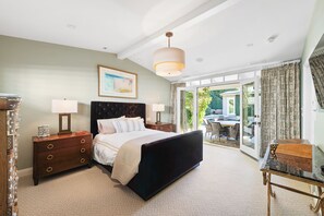 Ein Schlafzimmer mit einem Bett, Nachttischen, einer Kommode, einer Chandelüster und Blick ins Freie.