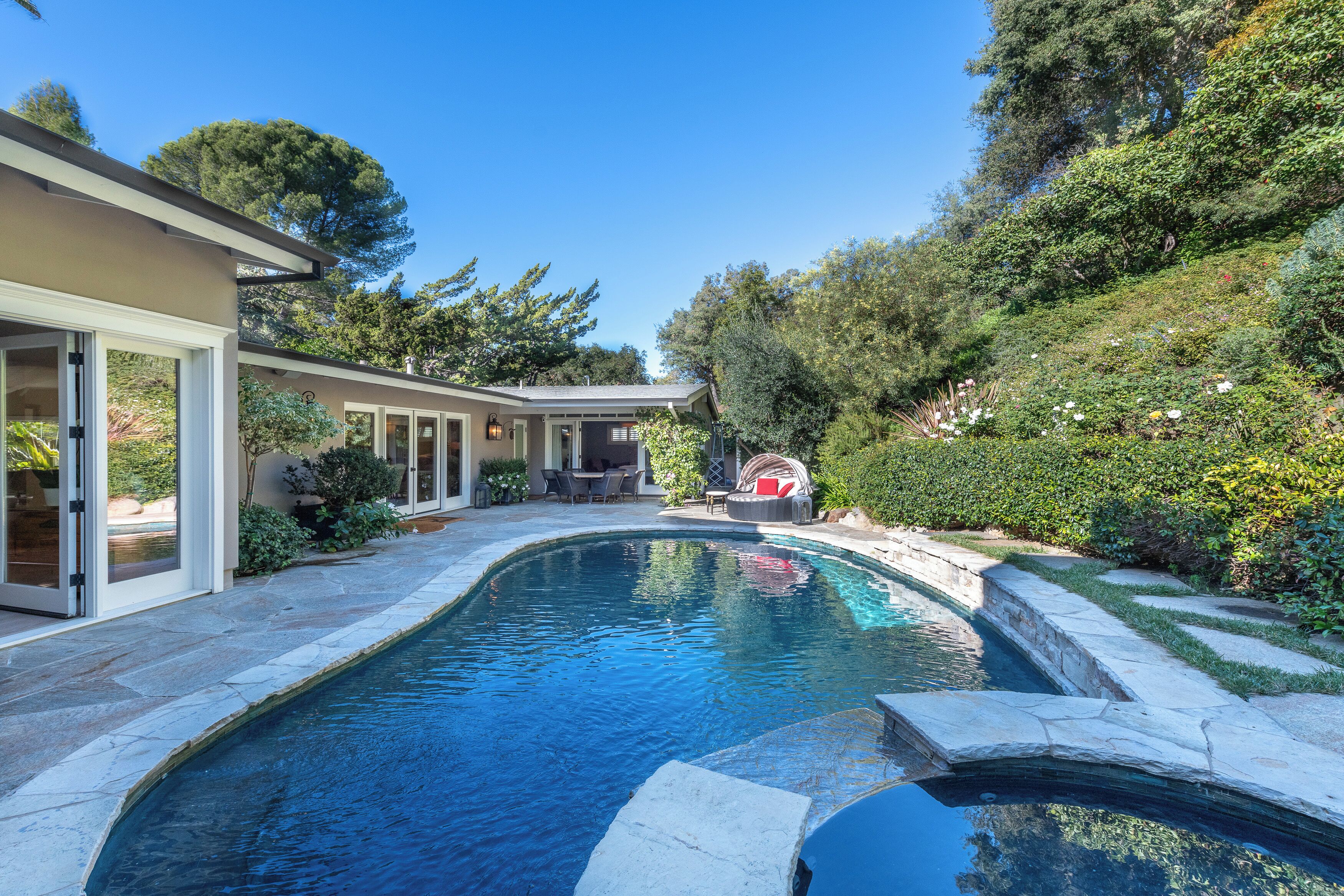 Une piscine avec un espace patio, un coin salon couvert et une maison dotée de portes-fenêtres coulissantes.