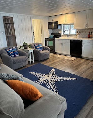 A living room with a star-patterned rug, grey sofas, and a kitchen with white cabinets and a black microwave.