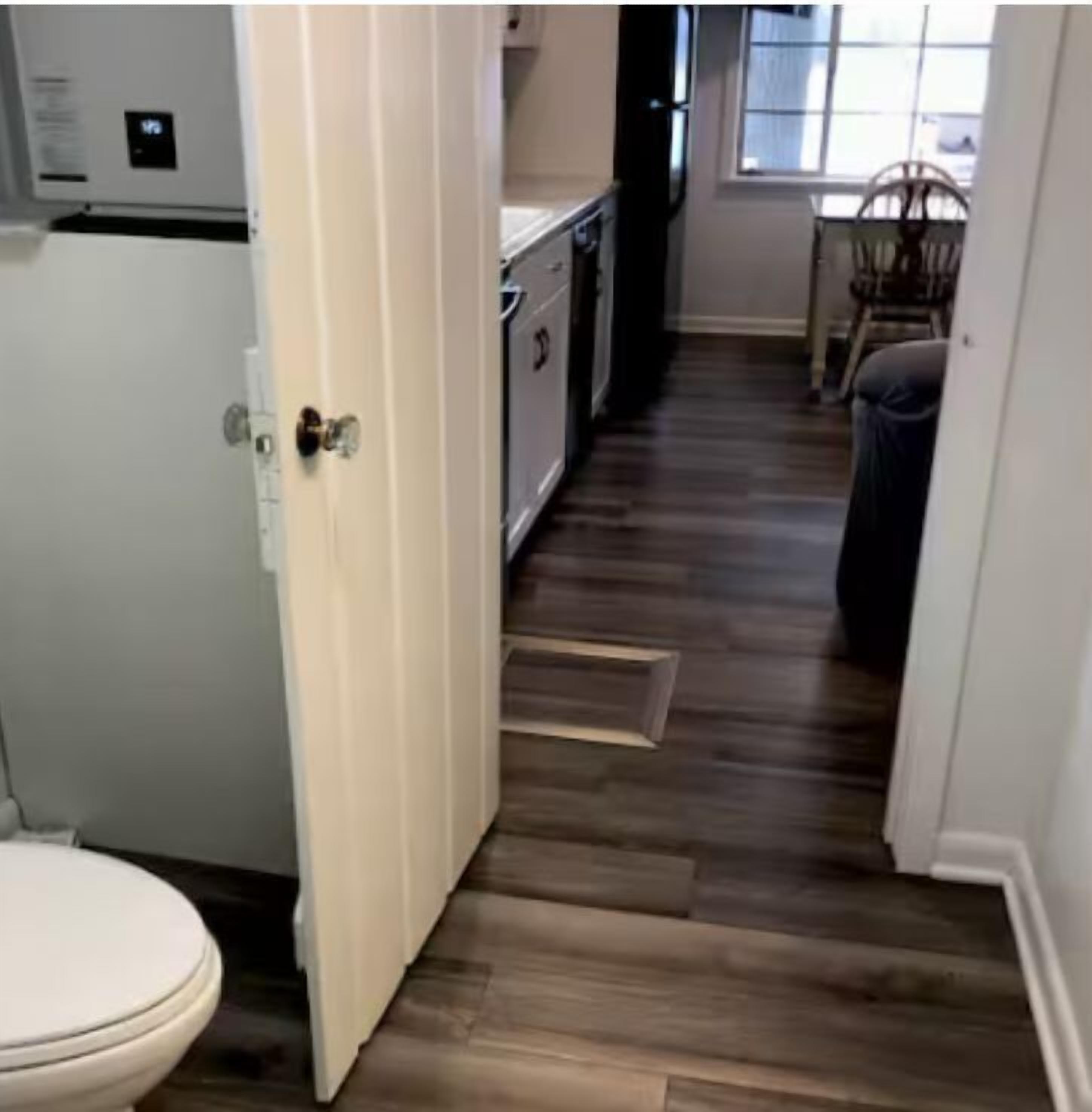 A narrow hallway with a wooden floor, a white door, a white toilet, and a glimpse of a kitchen area with cabinets and a window.