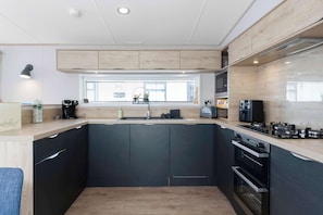 A modern kitchen with dark cabinets, a window, a built-in oven, and a countertop with a coffee maker. - Beachfront 2BR - Wi-Fi - Free Parking - Sleeps 6 (Southern Upland Way)