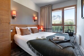 A hotel room with a bed, two bedside lamps, a window with curtains, a view of mountains, and a radiator. - Mountain View Retreat With Balcony for 8 (Araches-la-Frasse)