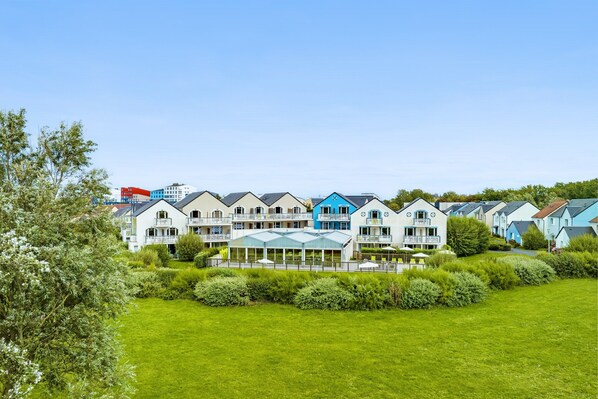 A row of modern, multi-story buildings with balconies, a covered outdoor area, and a well-maintained lawn. - Elegant 4-pax Apartment With River View (Courseulles-sur-Mer)