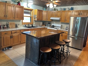 A kitchen with wooden cabinets, a stainless steel refrigerator, a large island with stools, and a ceiling fan. - Beautiful log cabin minutes away from Castle Rock lake & Buckhorn St. park (New Lisbon)
