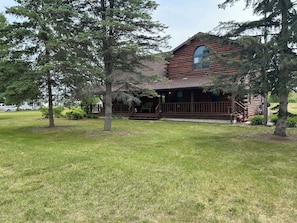 A log cabin with a porch, surrounded by trees and a grassy yard.