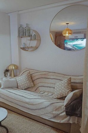 Un salon comprenant un canapé, un miroir rond, une étagère murale ornée d’objets décoratifs et un téléviseur diffusant un paysage pittoresque.