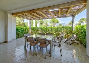 An outdoor patio with a glass-top table and chairs, surrounded by greenery and a wooden pergola. - Lecci Apartment With Outdoor Pool (Lecci)