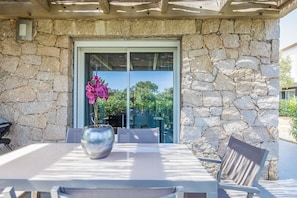A stone exterior wall, a glass door, a table with chairs, and a potted plant with pink flowers. - Lecci Apartment With Outdoor Pool (Lecci)