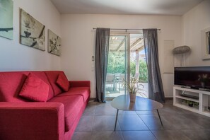 A living room with a red sofa, a glass table, a TV on a white stand, and a view through a glass door. - Lecci Apartment With Outdoor Pool (Lecci)