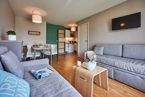 A modern living room with a grey sofa, a small wooden table with a teapot, and a dining area with white chairs and a table. - Courseulles Family Apartment for 6 (Courseulles-sur-Mer)