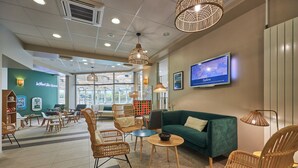 A hotel lobby with wicker furniture, a green sofa, a television, and a large window. - Courseulles Family Apartment for 6 (Courseulles-sur-Mer)