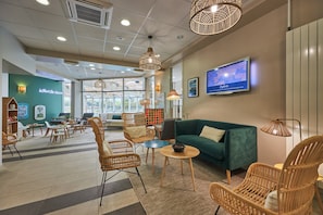 A hotel lobby with wicker furniture, a green sofa, a television, and a large window. - Courseulles Family Apartment for 6 (Courseulles-sur-Mer)