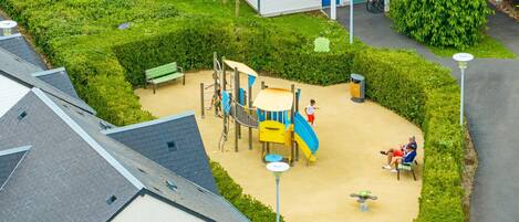 A playground with a slide and climbing structures, surrounded by a well-maintained lawn and a building with a balcony.
