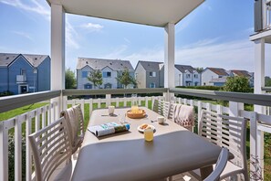 A covered outdoor patio with a table set for breakfast, white chairs, and a view of residential buildings. - Courseulles Family Apartment for 6 (Courseulles-sur-Mer)
