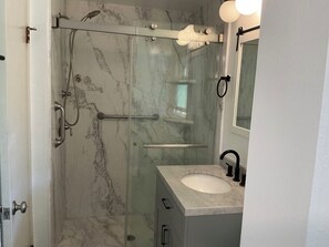 Ein Badezimmer mit einer gläsernen Duschkabine, einer Marmorwand, einem Waschbecken mit Spiegel und einer Tür.
