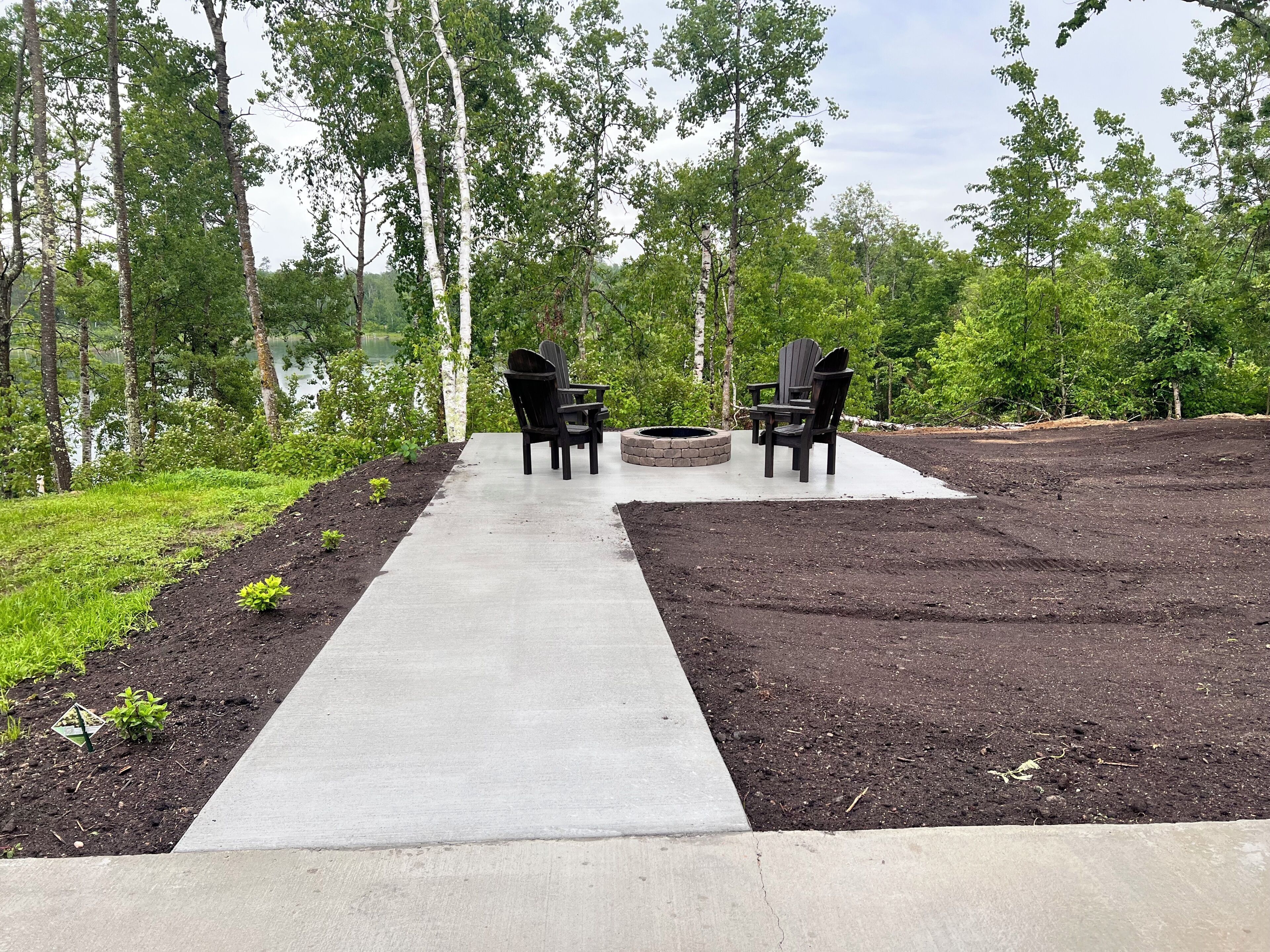 A concrete pathway leading to two Adirondack chairs with a fire pit in between, surrounded by a forested area.