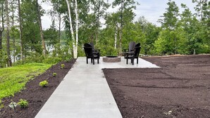 A concrete pathway leading to two Adirondack chairs with a fire pit in between, surrounded by a forested area. - New Lake Front Cabin 2BD 2Bath-Sofa bed - hot tub-18 miles to Itasca State Park (Bagley)